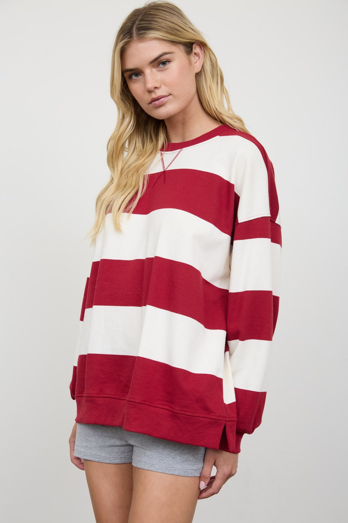 A woman modeling an oversized long-sleeve sweatshirt with bold horizontal red and off-white stripes, paired with gray shorts, against a light gray studio background.