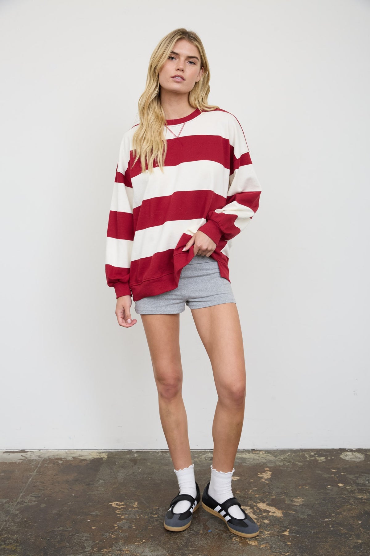 A woman modeling a red and white horizontally striped long-sleeve crewneck sweatshirt, paired with gray shorts, white socks, and black sandals, standing against a white wall.