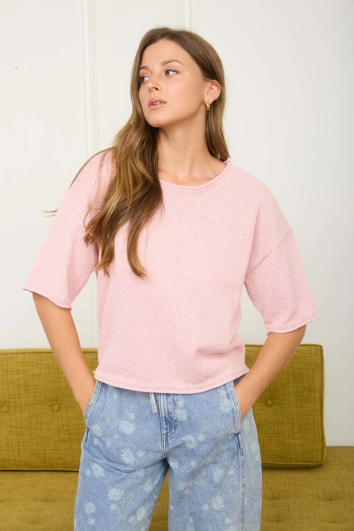 Woman wearing a pink oversized shirt and blue jeans standing against a white wall.
