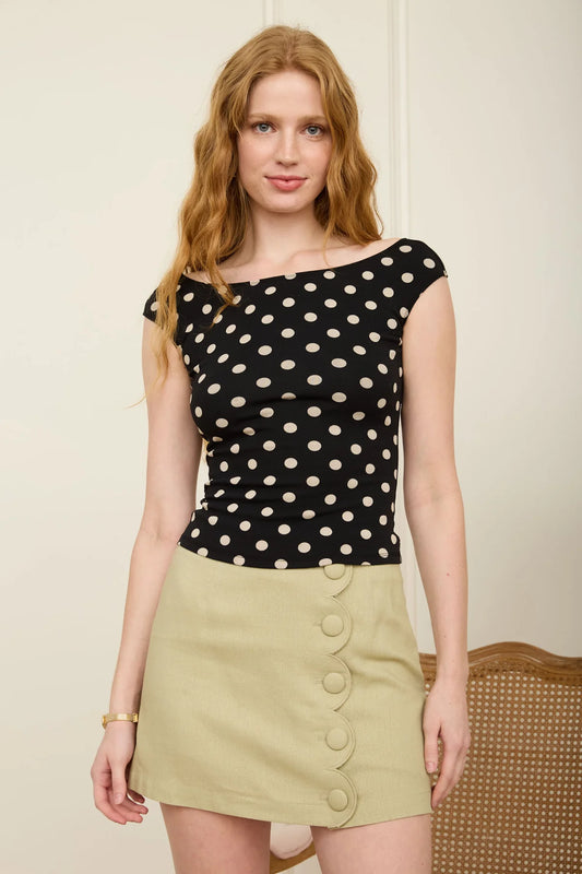 Woman wearing a black polka dot top and beige skirt against a plain background