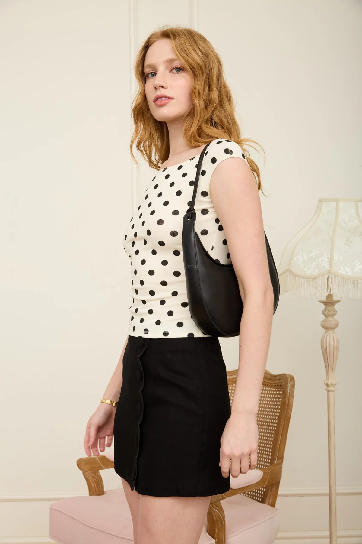Woman wearing a polka dot top and black skirt holding a black handbag indoors.