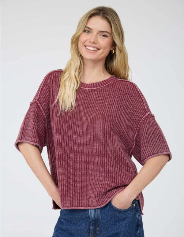 A woman wearing a ribbed short-sleeve acid washed burgundy sweater, paired with blue jeans, against a plain light gray studio background.