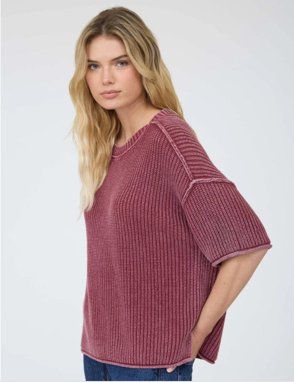 A woman modeling a ribbed knit short-sleeve acid washed burgundy sweater, paired with blue jeans, against a plain light gray studio background.