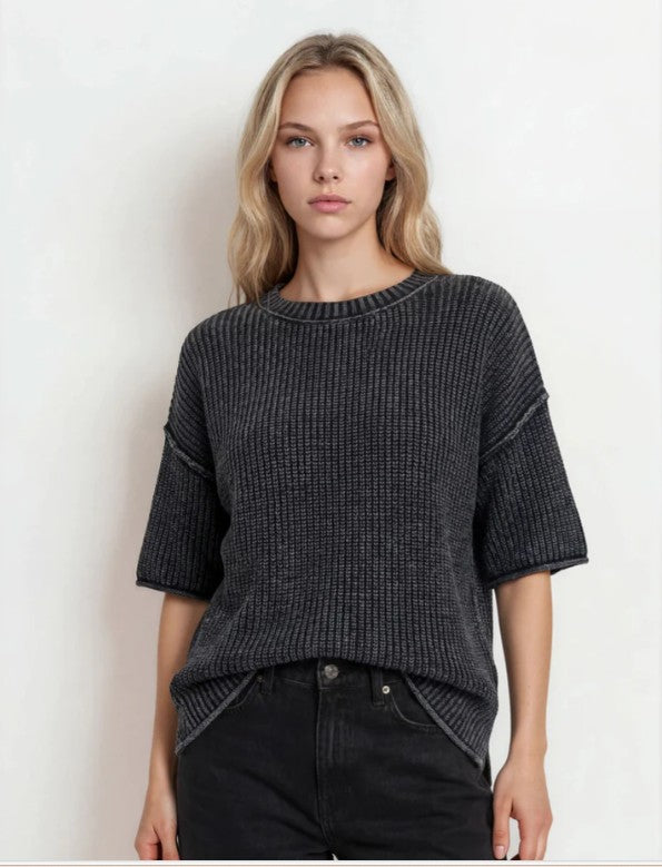 A woman wearing a dark charcoal gray ribbed knit short-sleeve sweater and black jeans, standing against a plain off-white background.