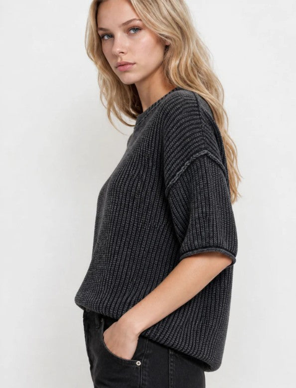 A woman wearing a dark charcoal gray ribbed knit short-sleeve top, shown from a side profile against a plain light gray background.