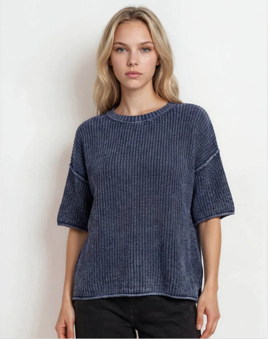 A woman wearing a textured, short-sleeve knit top in a dark navy blue color, paired with black pants, standing against a plain white background.