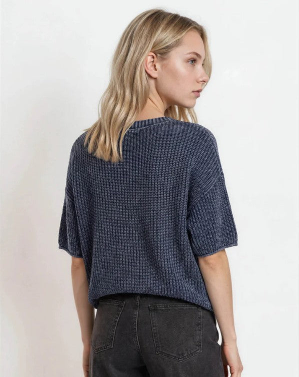 A woman with blonde hair is shown from the back wearing a navy blue ribbed knit short-sleeve top and dark gray jeans against a plain white background.