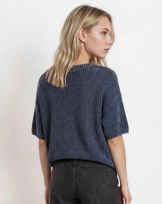 A woman with blonde hair is shown from the back wearing a navy blue ribbed knit short-sleeve top and dark gray jeans against a plain white background.