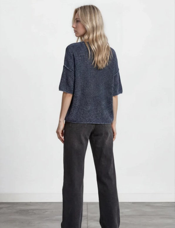 A woman shown from the back wearing a dark blue-gray textured short-sleeve knit top and dark charcoal wide-leg pants, standing against a plain white wall.