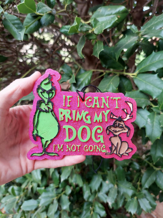 A hand holding a pink air freshener featuring the Grinch with a dog. The air freshener is shaped like a character from a Christmas themed story, with green details on a pink background, and it's set against a backdrop of greenery.