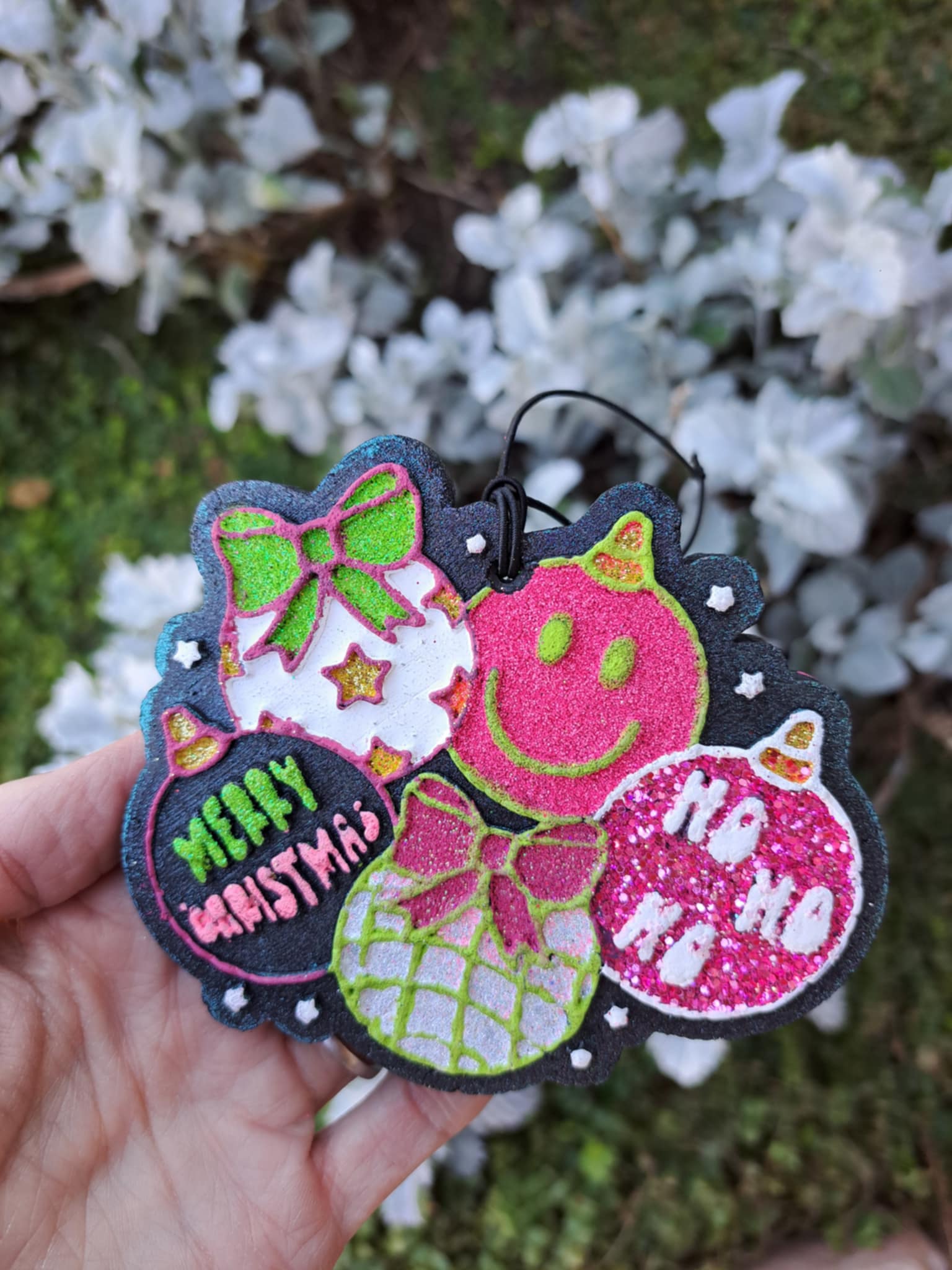 Decorative Christmas ornament freshies with a smiley face and text held against a blurred natural background.