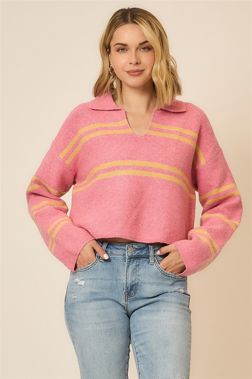 Woman wearing a pink vneck sweater with orange stripes and distressed jeans on a neutral background