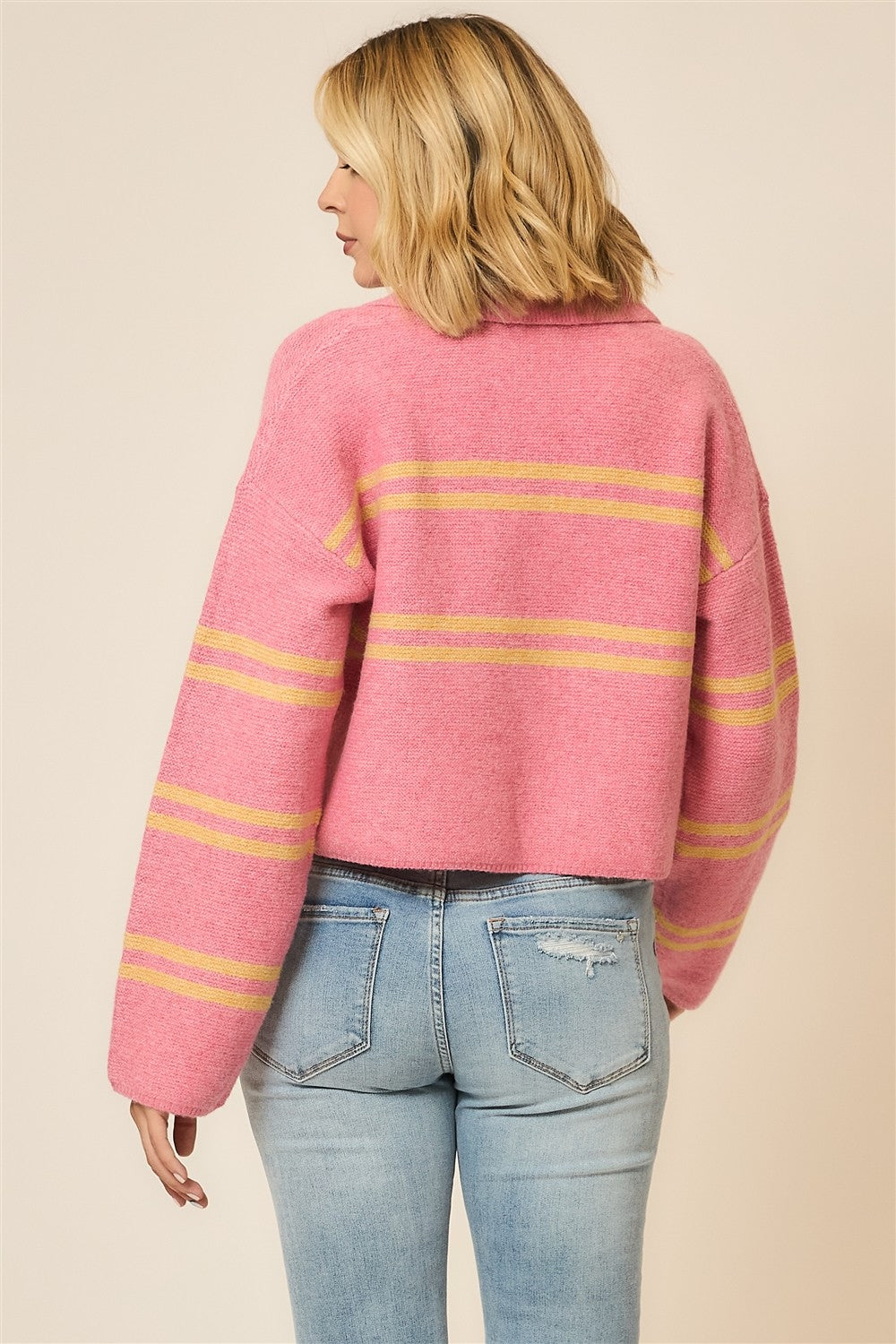 Back view of a woman wearing a pink V-neck sweater with orange stripes and distressed jeans on a neutral background
