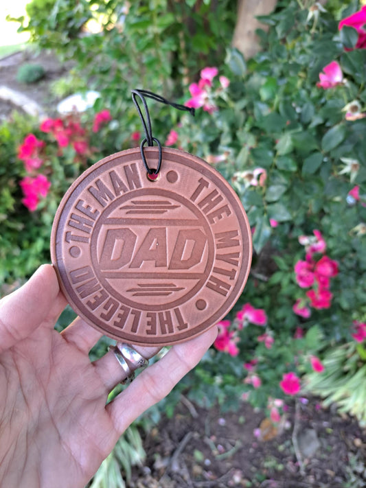A hand holding a round, brown leather freshie with the text 'DAD' in the center, surrounded by a blurred background of flowers.