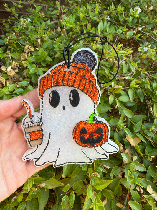 A hand holds a white felt ghost Halloween ornament with an orange knit hat, holding a jack-o'-lantern and a drink, set against a background of green garden foliage.