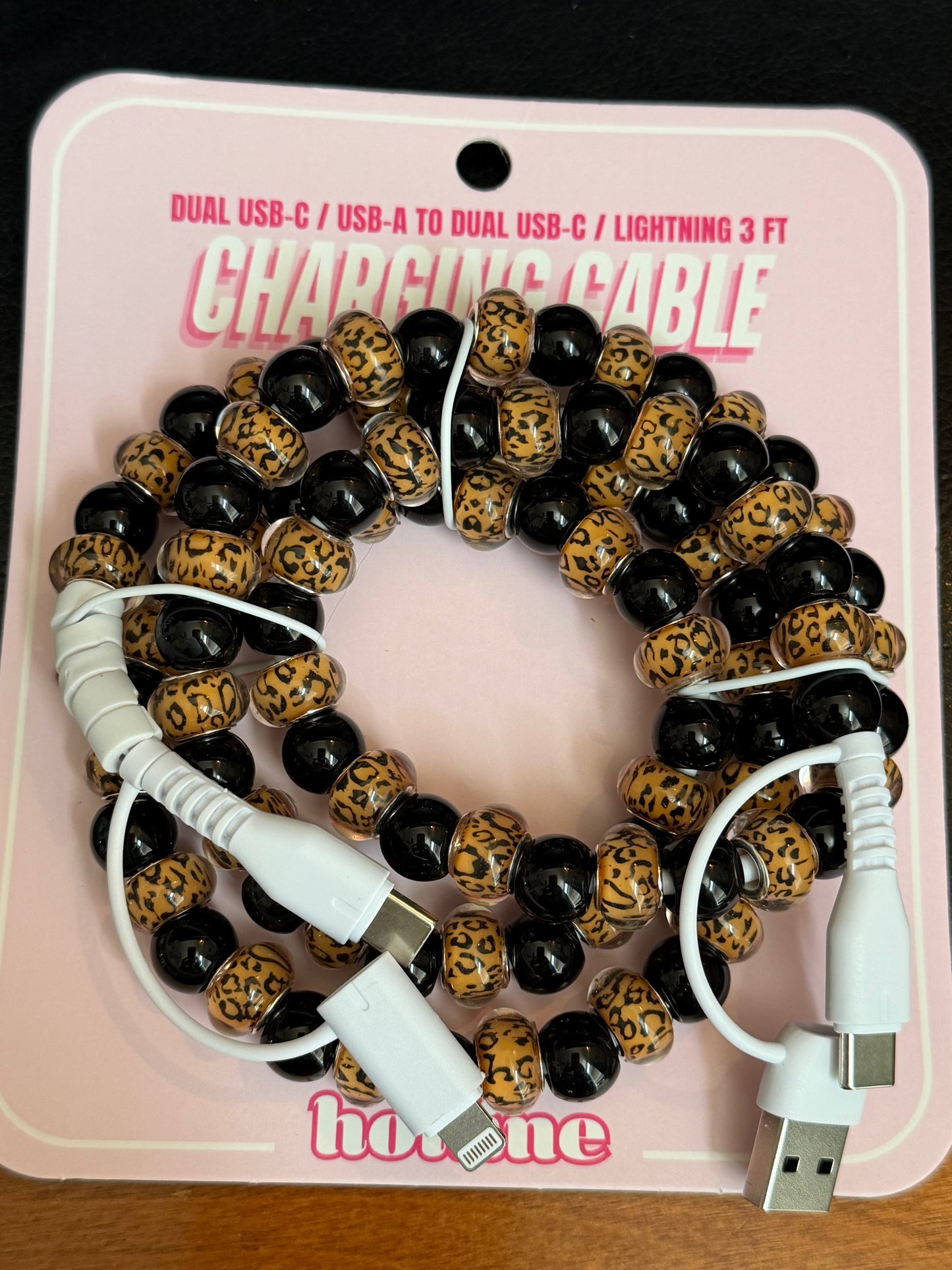 A beaded charging cable coiled on its pink retail backing card. The cable features alternating glossy black beads and leopard-print beads in tan and black, with white USB-C to USB-A and Lightning connectors visible at the ends.