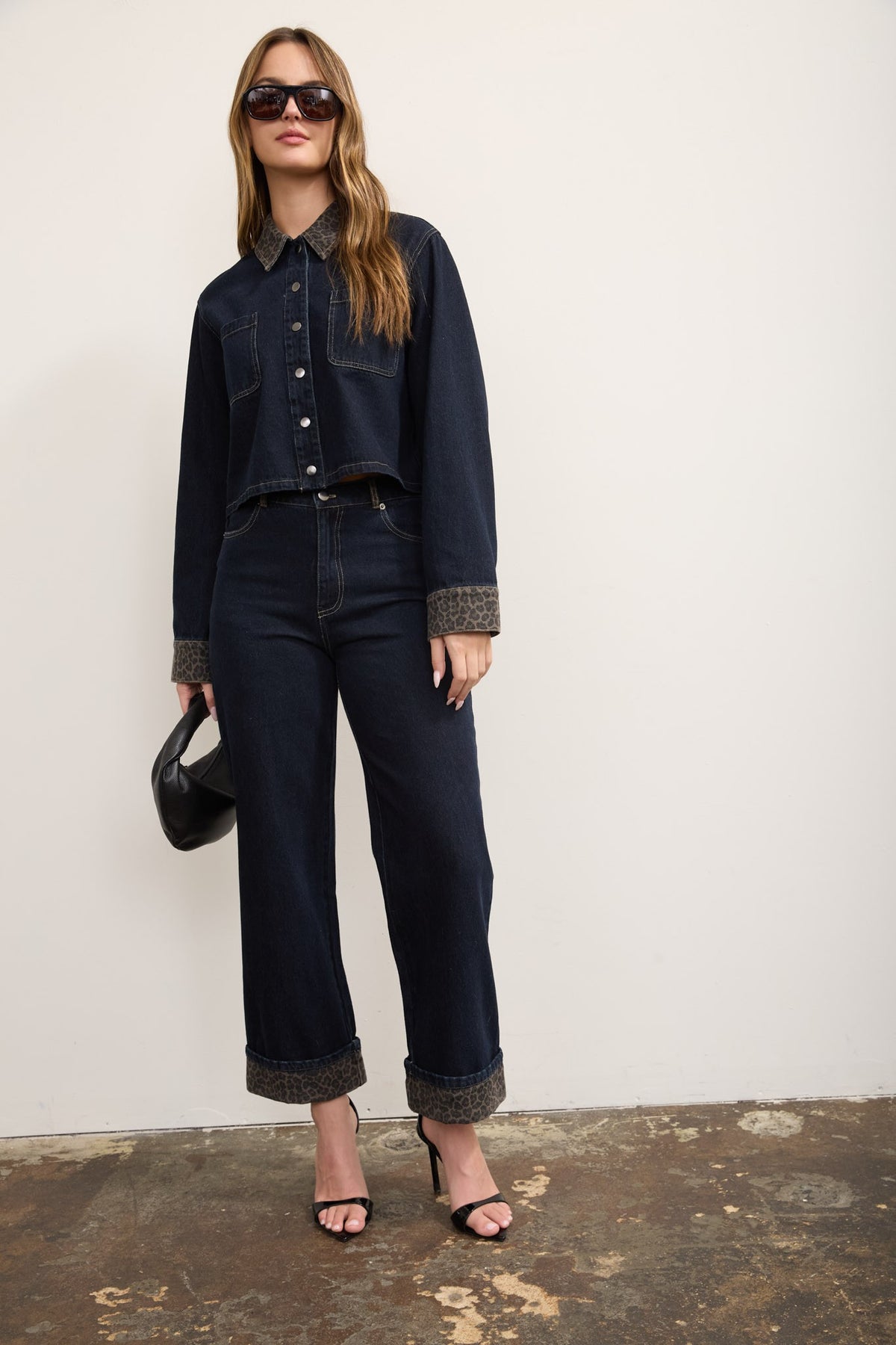 A model wearing a dark wash denim jacket and matching wide-leg jeans with contrasting brown textured cuffs and hem, styled with black sunglasses, a black handbag, and black heeled sandals against a plain off-white wall.