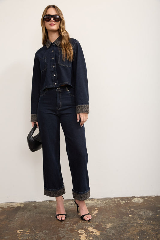 A model wearing a dark wash denim jacket and matching wide-leg jeans with contrasting brown textured cuffs and hem, styled with black sunglasses, a black handbag, and black heeled sandals against a plain off-white wall.