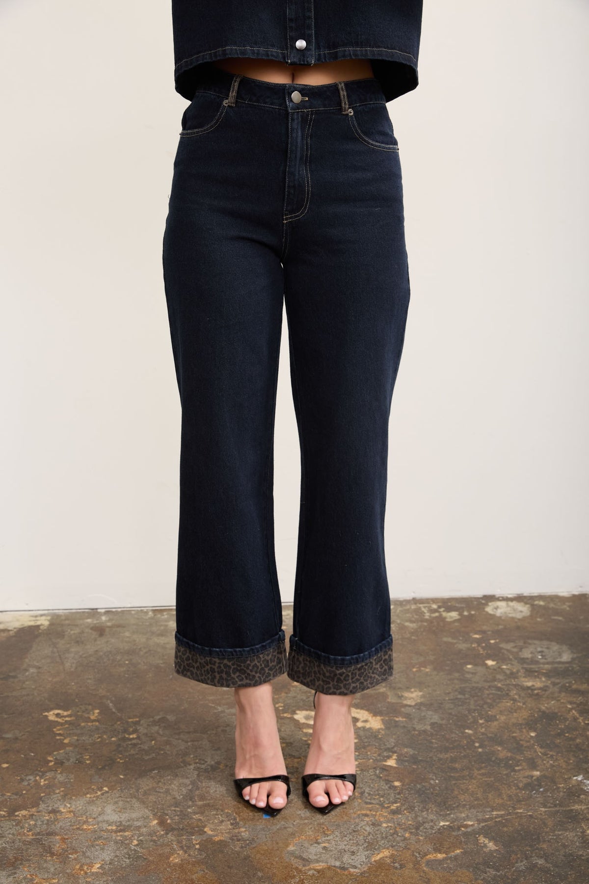 A person wearing dark cropped wide leg jeans with leopard print cuffs, paired with black heeled sandals against a plain light wall and rustic floor.