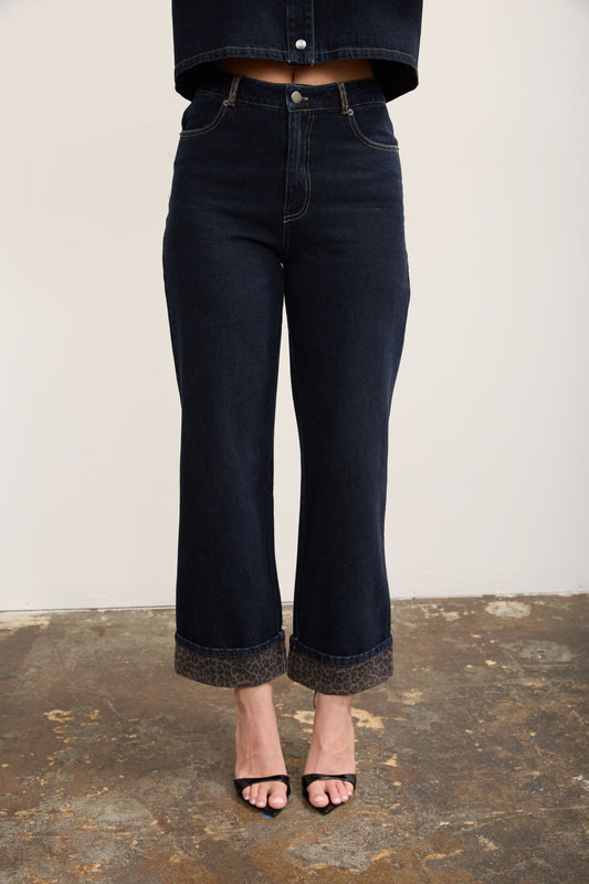 A person wearing dark cropped wide leg jeans with leopard print cuffs, paired with black heeled sandals against a plain light wall and rustic floor.