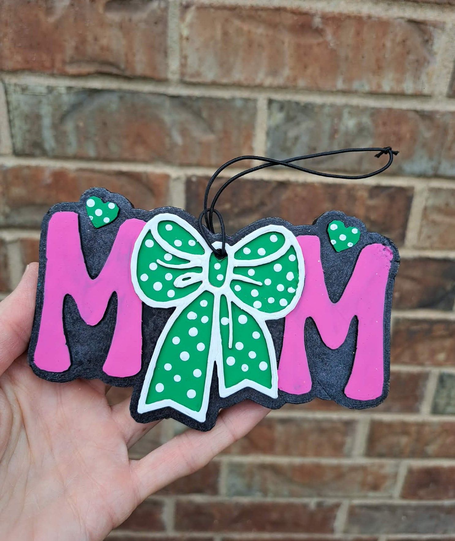 Decorative 'MOM' freshie with a green polka dot bow held against a brick wall.
