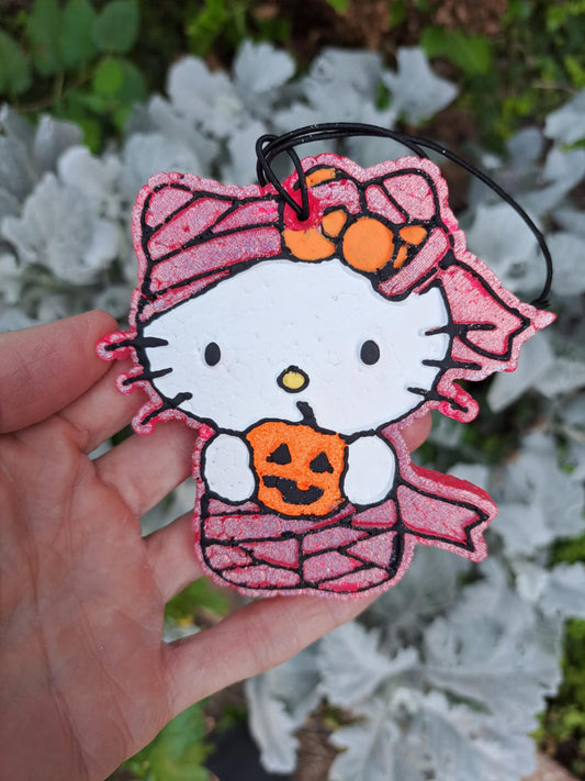 Mummy kitty holding pumpkin air freshener held by a hand with a blurred natural background