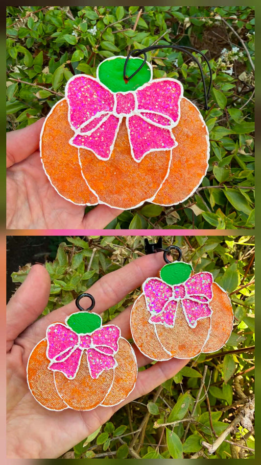 A hand holds two pumpkin-shaped decorative freshies outdoors against green foliage. Each ornament is orange with a glittery pink bow and a green top, outlined in white, with black hanging loops.