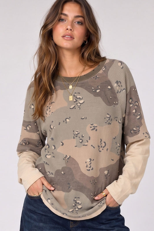 A woman wearing a long-sleeve camouflage knit top with beige sleeves and scattered rhinestone embellishments, paired with dark blue jeans, against a light gray studio background.