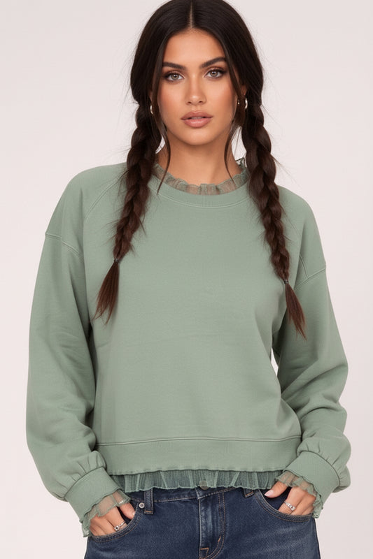 A woman with dark braids wears a sage green crewneck sweatshirt with ruffled trim at the collar and hem, paired with dark blue jeans, against a plain light background.