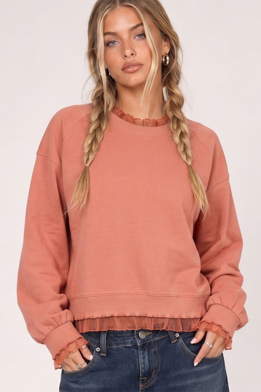 A woman with blonde braids wears a dusty rose crewneck sweatshirt with ruffled trim at the collar and hem, paired with dark blue jeans, against a plain light background.
