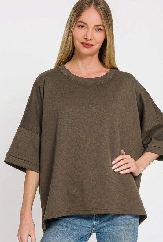 A woman wearing an oversized olive brown short-sleeve top with three-quarter sleeves and a relaxed fit, paired with blue jeans, against a plain white background.