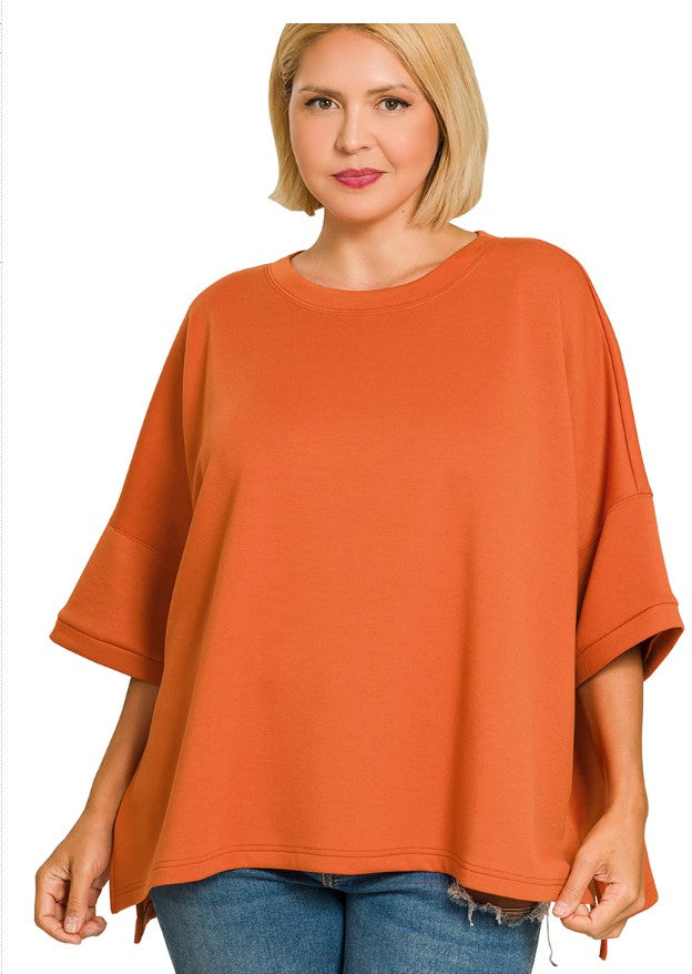 A woman wearing a loose-fitting, burnt orange short-sleeve top with raw hems and side slits, paired with blue jeans, against a plain white background.