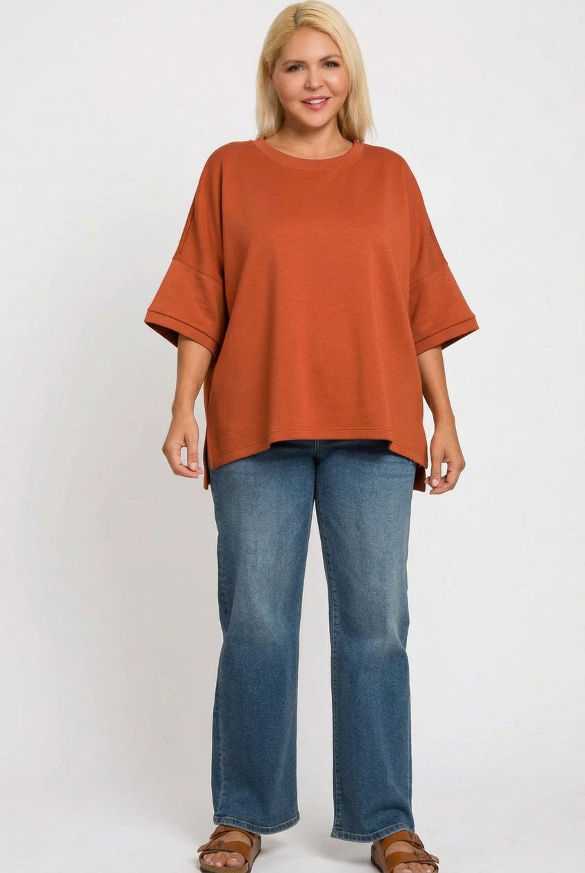 A woman modeling a dark charcoal gray oversized short-sleeve top with blue jeans and brown sandals, standing against a plain white background.
