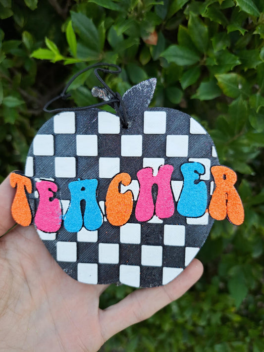 Checkered apple shaped freshie with 'teacher' across it, held against a green leafy background.