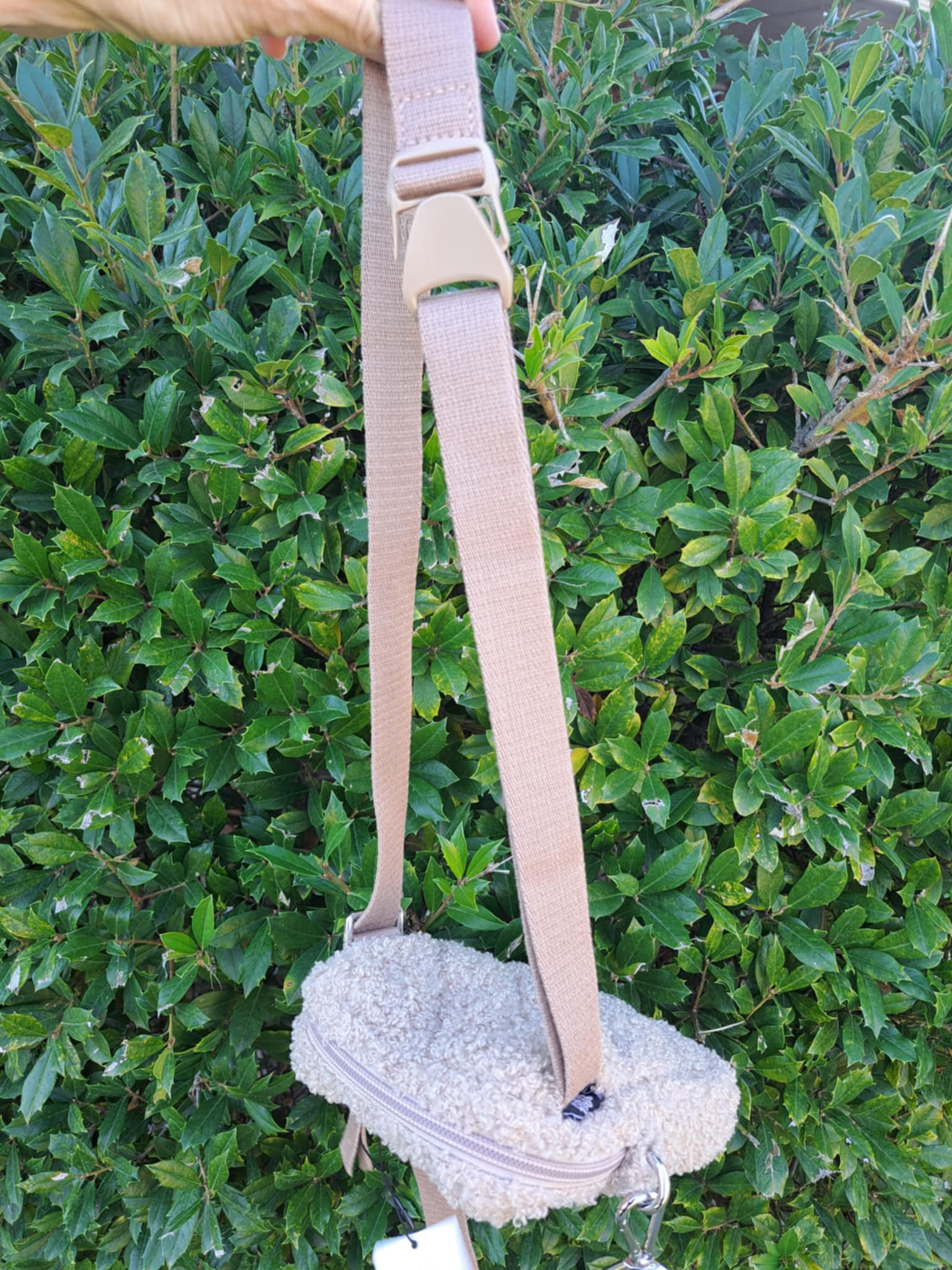 Small white bag with a beige belt strap against a green bush background