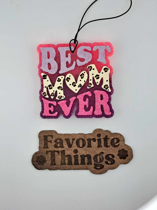 A handmade hanging air freshener with 'BEST MOM EVER' painted in leopard print design, with pink and purple ombre shades on a black background. Below it, the phrase 'Favorite Things' is written in a simple font.