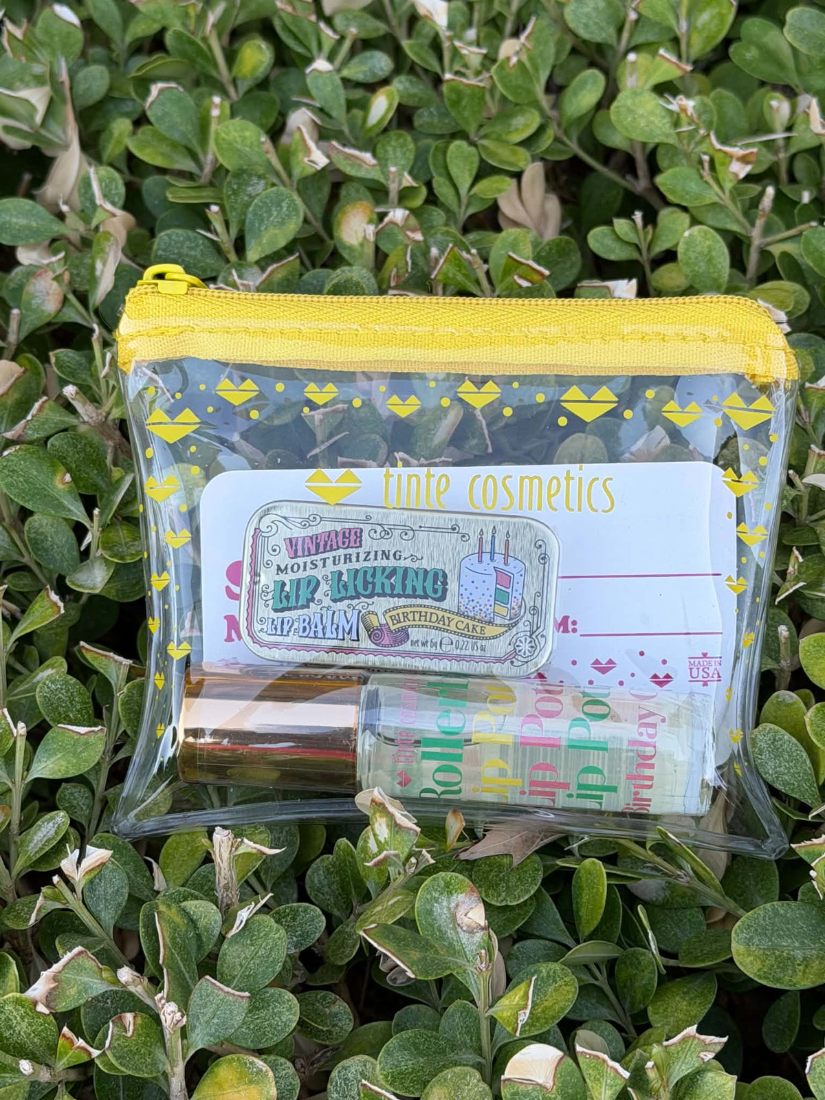 A clear plastic zip pouch with a bright yellow zipper and yellow heart pattern, containing lip balm and a card labeled 'Lime cosmetics', placed on green foliage.