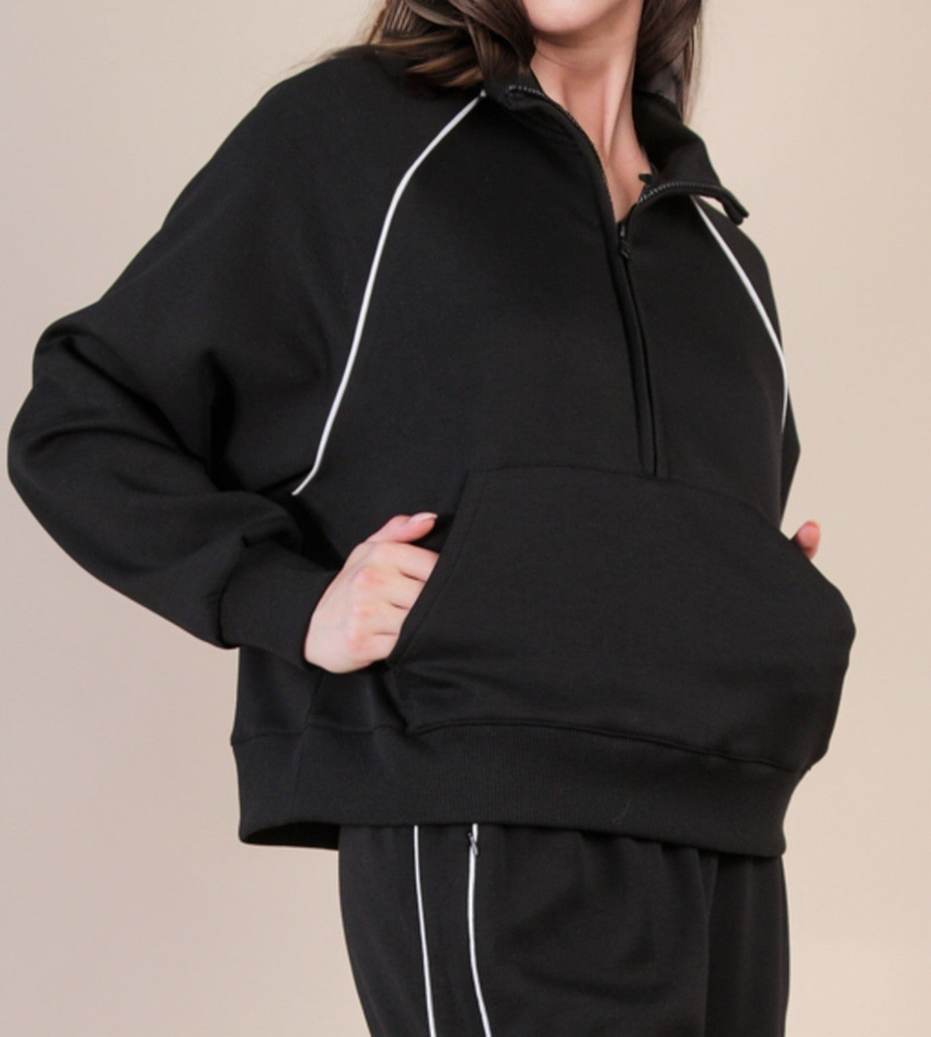 Black jogging top with pockets and white piping on a woman. Beige background.