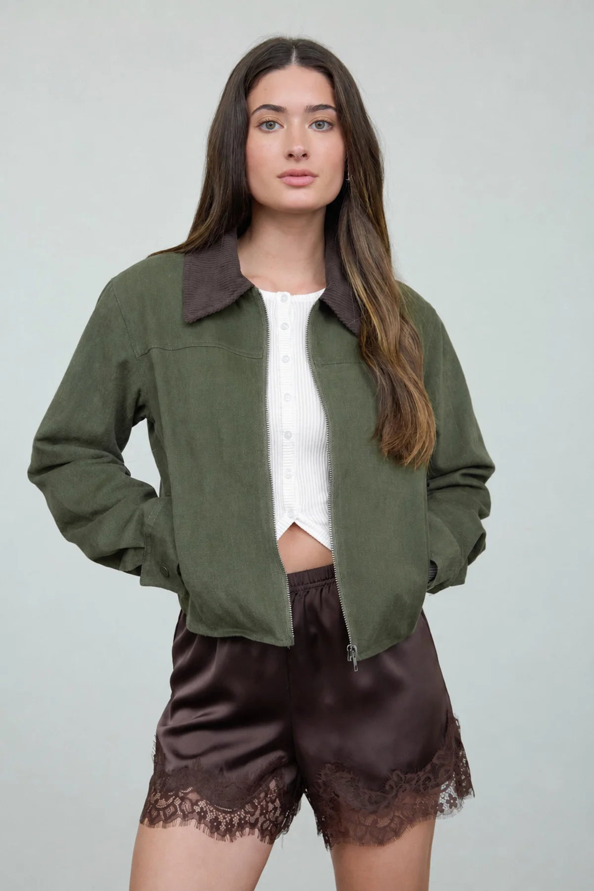 A woman modeling an olive green zip-up jacket with a dark brown collar, worn over a white cropped top and dark brown satin shorts with lace trim, against a light gray studio background.