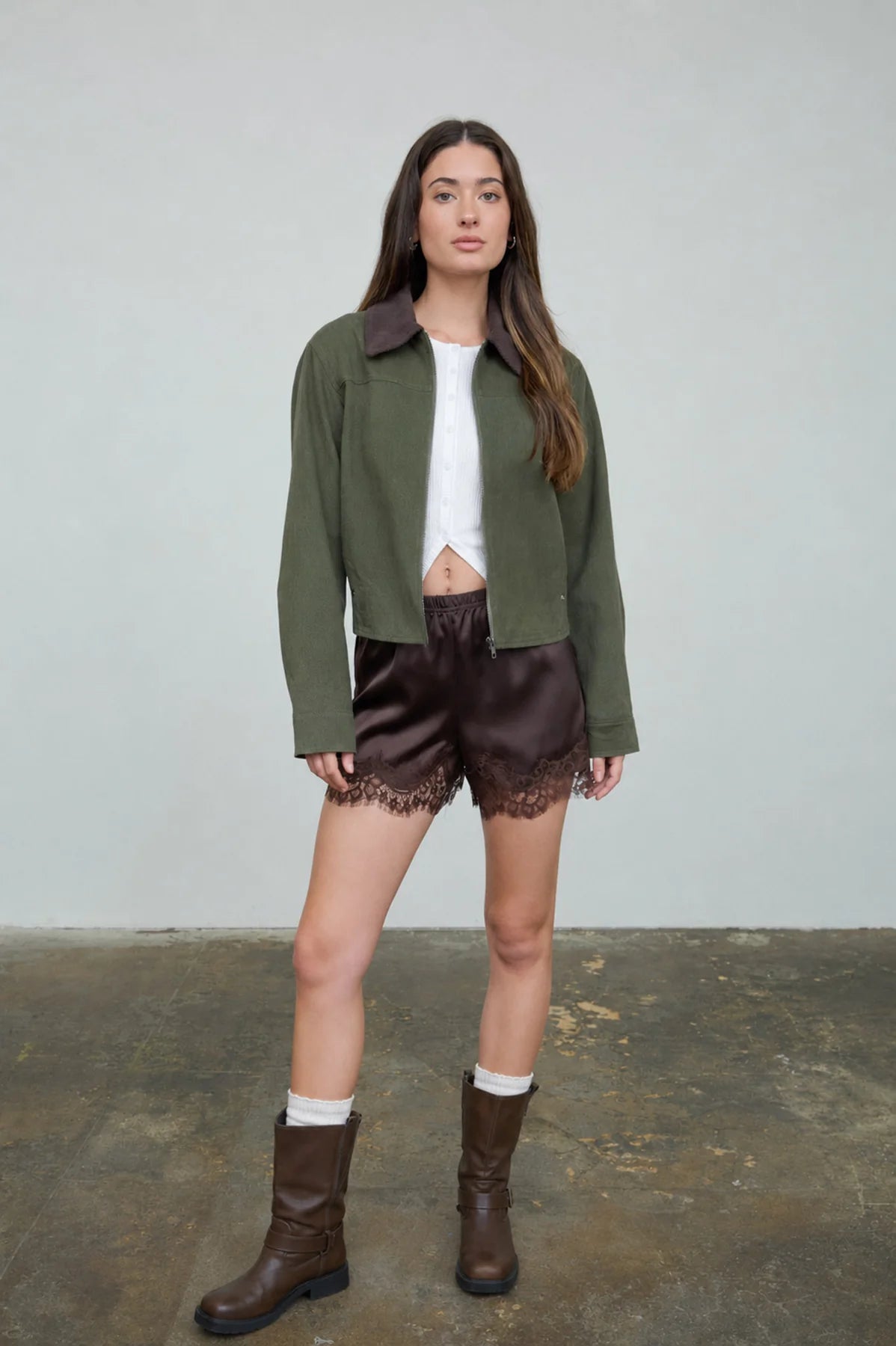A woman modeling a cropped olive green jacket with dark brown lapels, worn over a white crop top and paired with dark brown satin shorts featuring a lace hem, brown socks, and brown boots, set against a plain light gray wall and concrete floor.