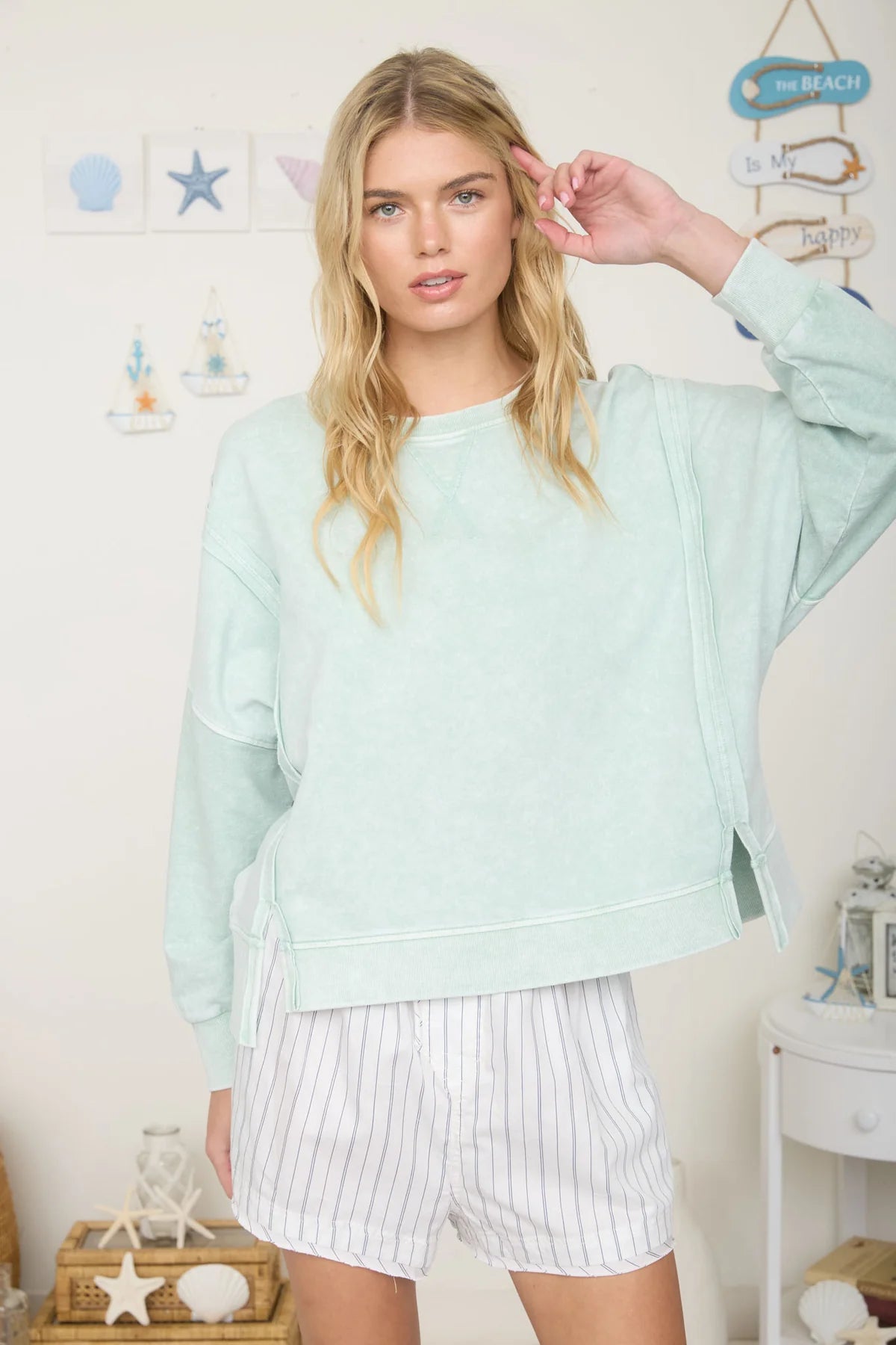 Woman wearing a light green sweatshirt and white shorts in a room with decorative items on the wall.