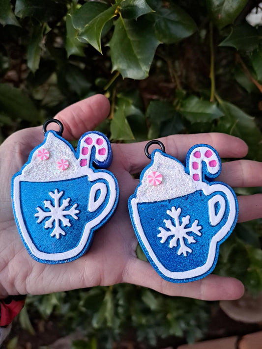Two blue metallic air vent clips shaped like mugs with snowflake patterns, held in a person's hands. Green background.