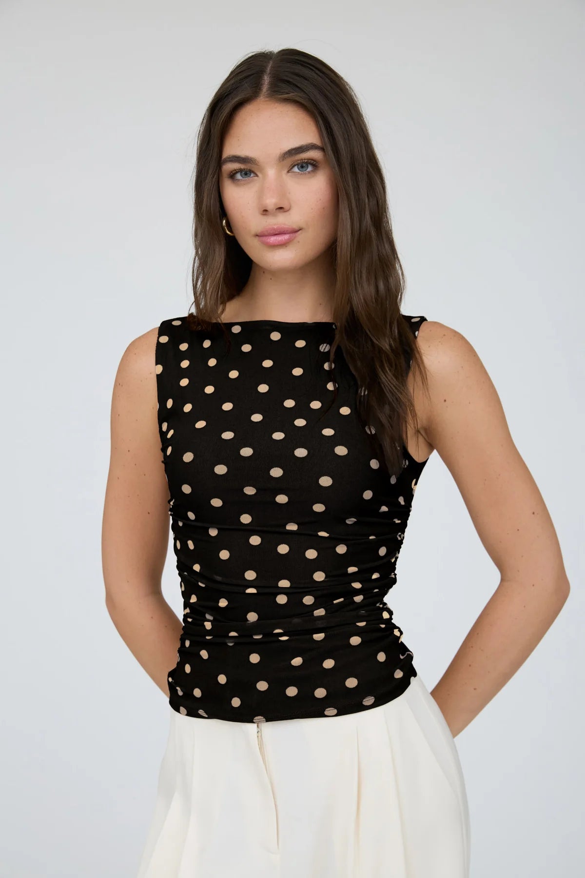 A woman modeling a sleeveless black top with beige polka dots and gathered detailing at the waist, paired with white bottoms, against a light gray studio background.