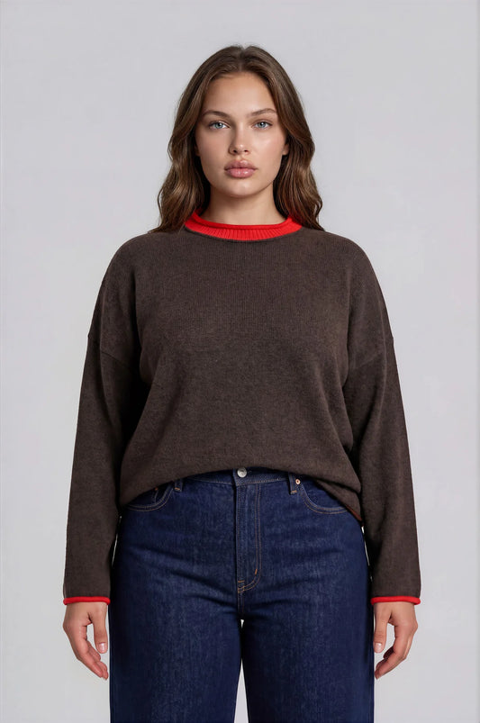 A woman modeling a dark brown long-sleeve knit sweater with a contrasting red crew neck and red cuffs, paired with dark blue jeans, against a light gray studio background.