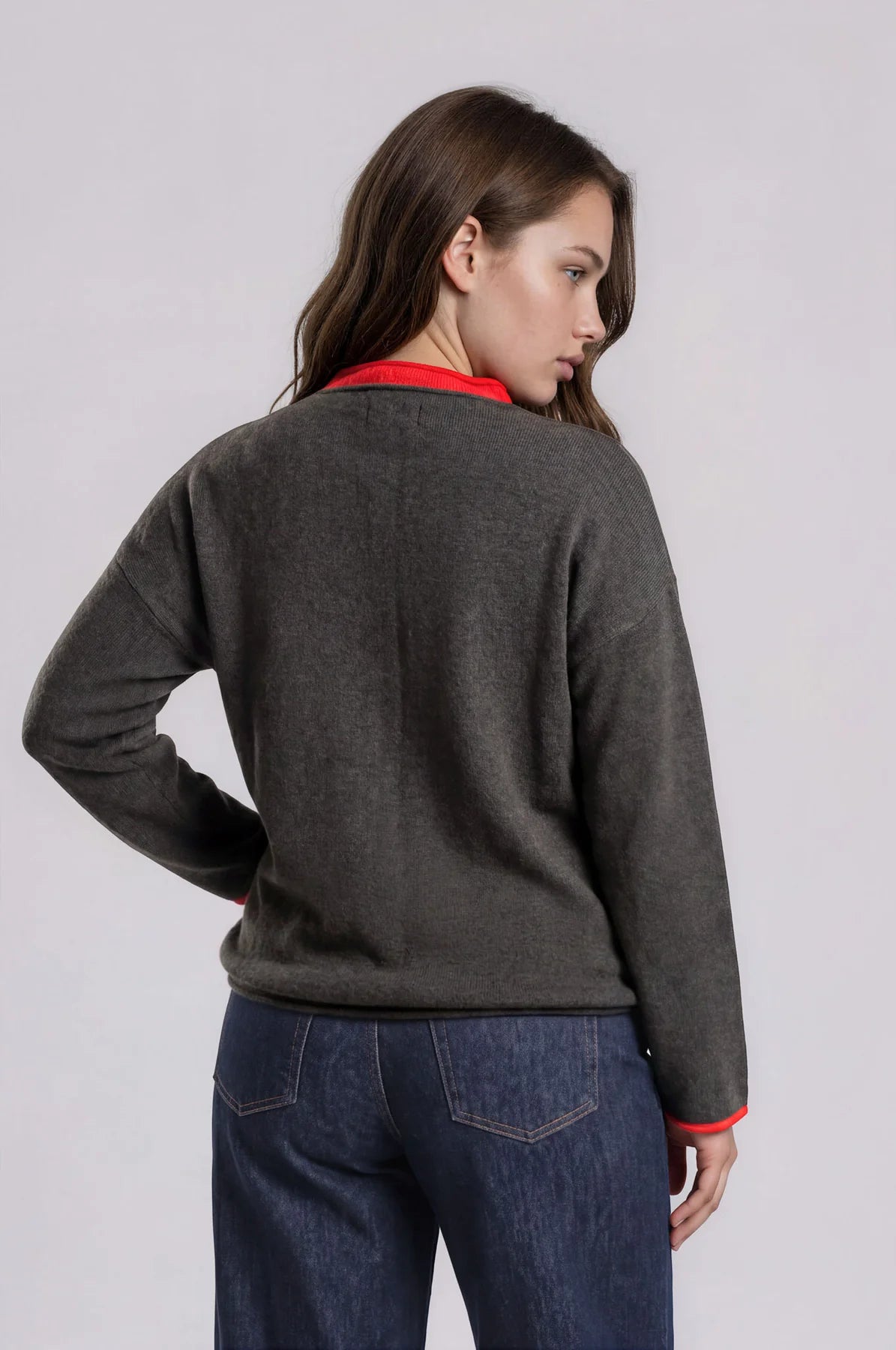 A woman shown from the back wearing a dark gray knit sweater with bright red ribbed trim at the collar and cuffs, paired with dark blue jeans, against a light gray studio background.