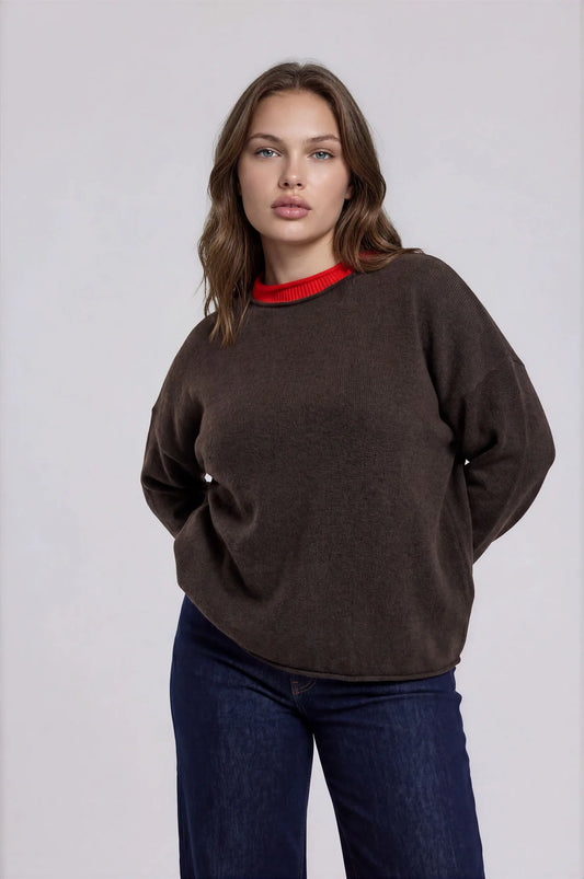 A woman modeling an oversized dark brown crewneck sweater with a contrasting red inner collar, paired with dark blue jeans, against a plain light gray studio background.