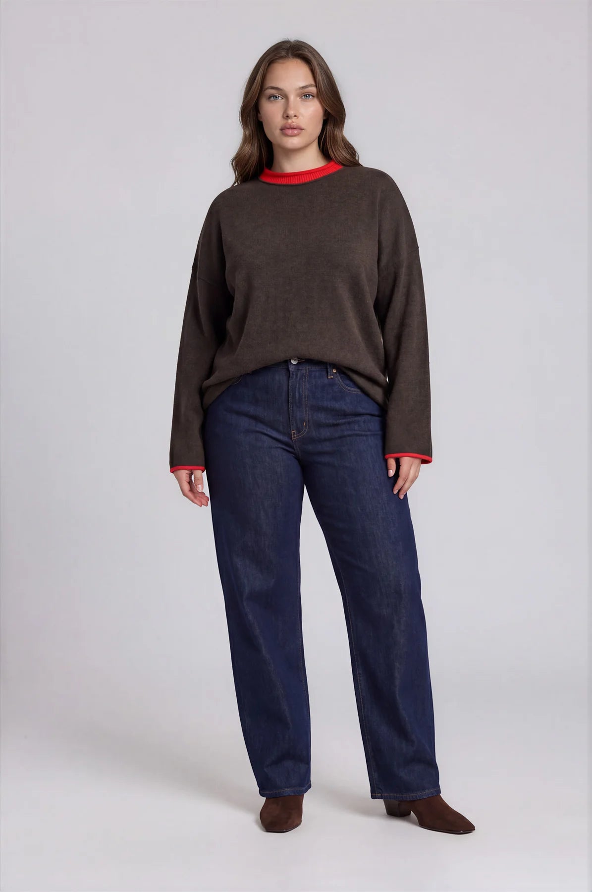 A woman modeling a dark brown crewneck sweater with red trim and dark blue jeans, standing against a light gray studio background.