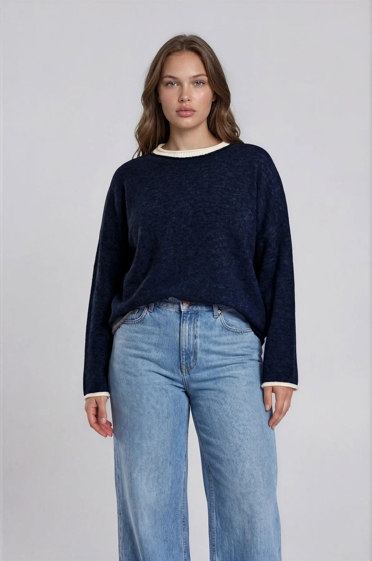 A woman modeling a dark navy blue knit sweater with contrasting cream ribbed trim at the crew neck and cuffs, paired with light blue wide-leg jeans, against a neutral light gray background.