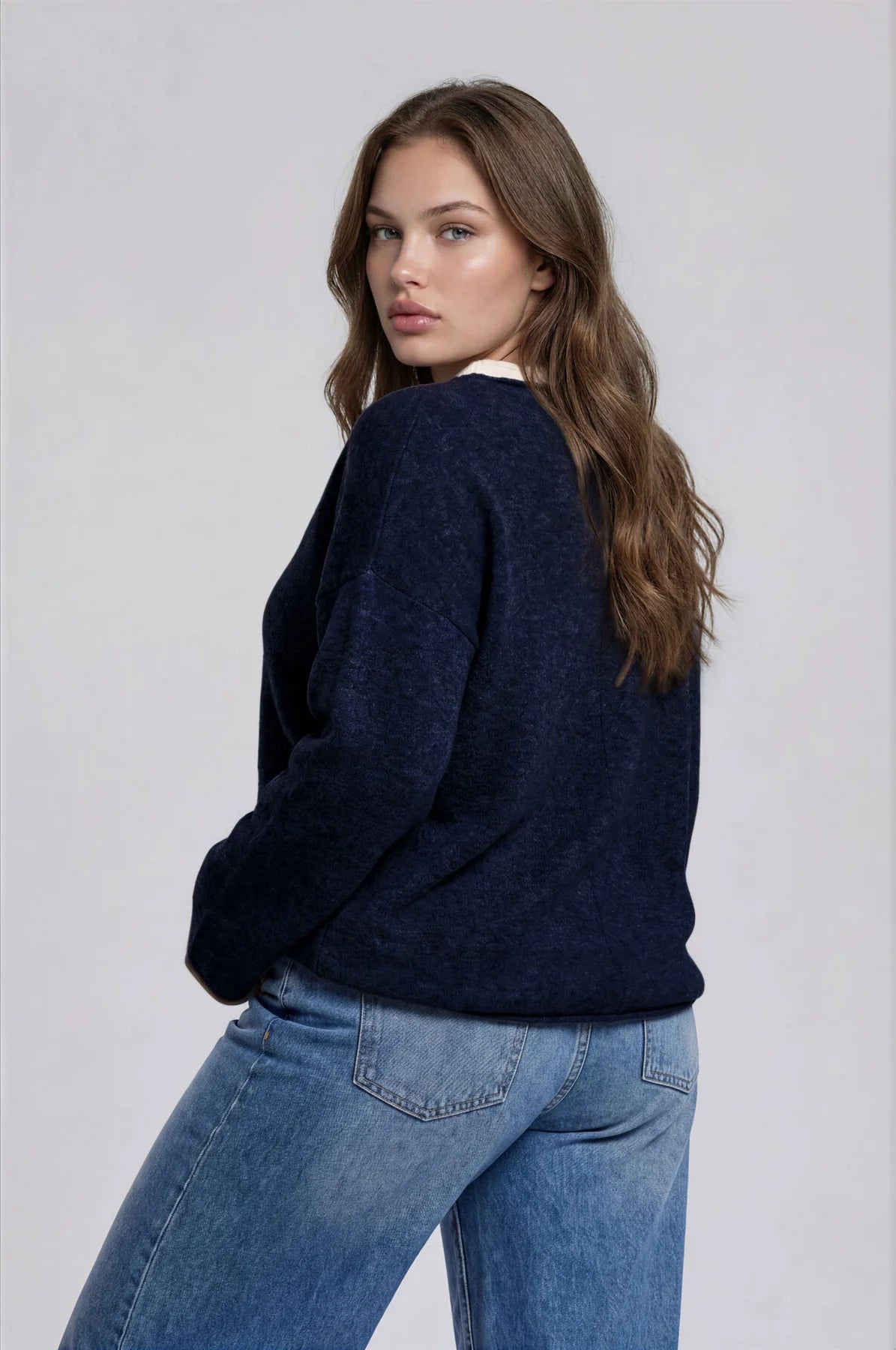 A woman wearing a dark navy blue knit sweater and light blue jeans, photographed from the back against a plain light gray studio background.