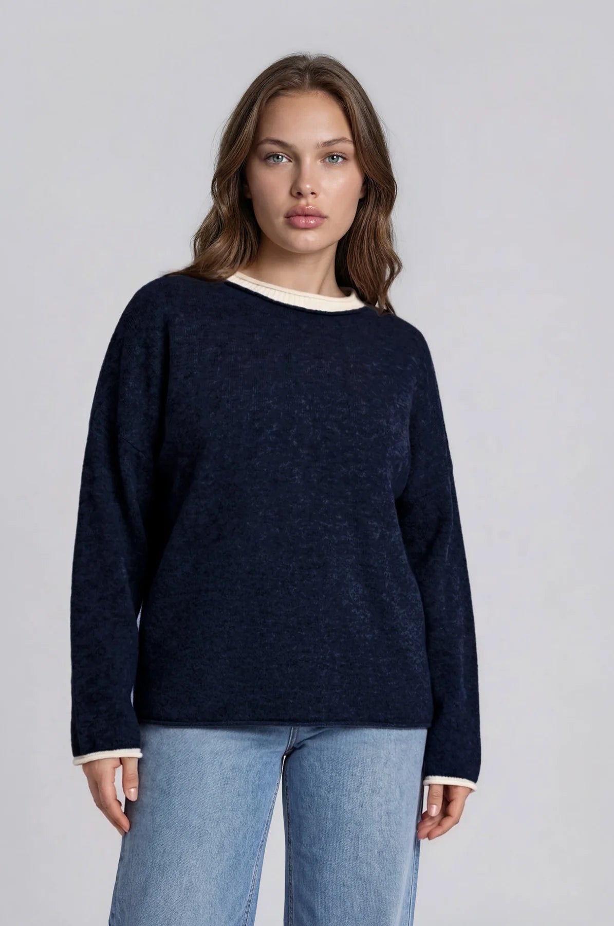 A woman modeling a dark navy blue knit sweater with contrasting cream ribbed trim at the crew neckline and cuffs, paired with light blue jeans, against a light gray studio background.